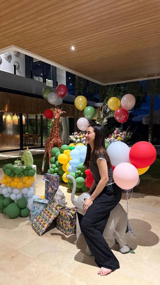 Seorang wanita tersenyum duduk di patung gajah kecil di pesta ulang tahun anak dengan dekorasi balon warna-warni dan hadiah di sekitarnya.