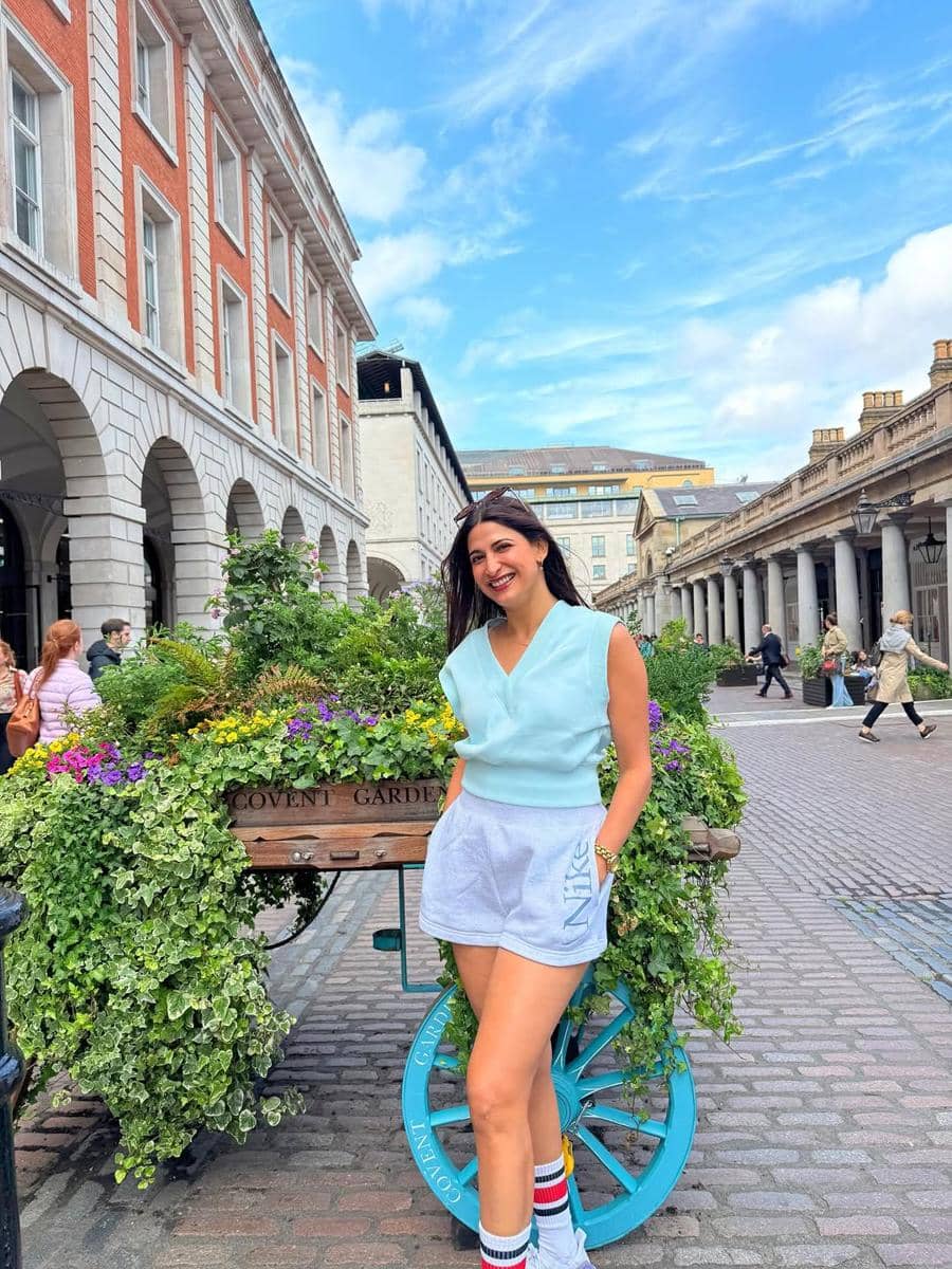 Seorang wanita tersenyum mengenakan atasan biru muda dan celana pendek abu-abu berpose di depan gerobak bunga di Covent Garden, London.