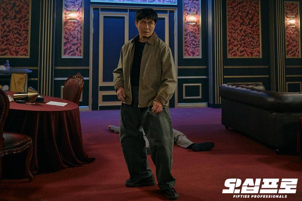 Seorang pria berdiri di ruangan bergaya klasik dengan karpet merah dan dinding berpola, sementara seseorang tergeletak di lantai di belakangnya.
