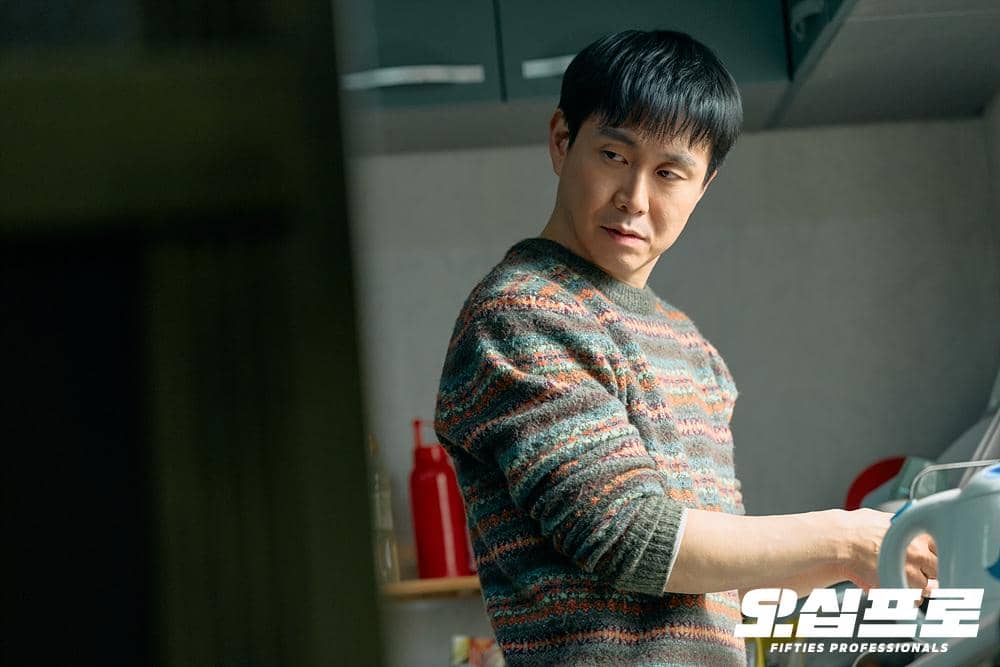 Seorang pria mengenakan sweter bermotif garis sedang berada di dapur sambil menatap ke samping dengan ekspresi serius.