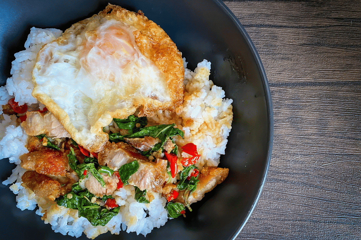 Semangkuk nasi putih dengan telur goreng di atasnya, disajikan bersama potongan daging, cabai merah, dan sayuran hijau di atas meja kayu.