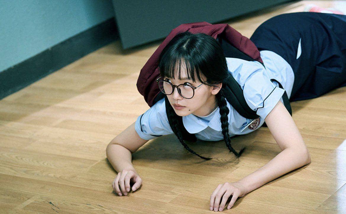 Seorang siswi berkacamata dengan seragam sekolah dan ransel merah merangkak di lantai kayu dalam cuplikan drama Teach You A Lesson.