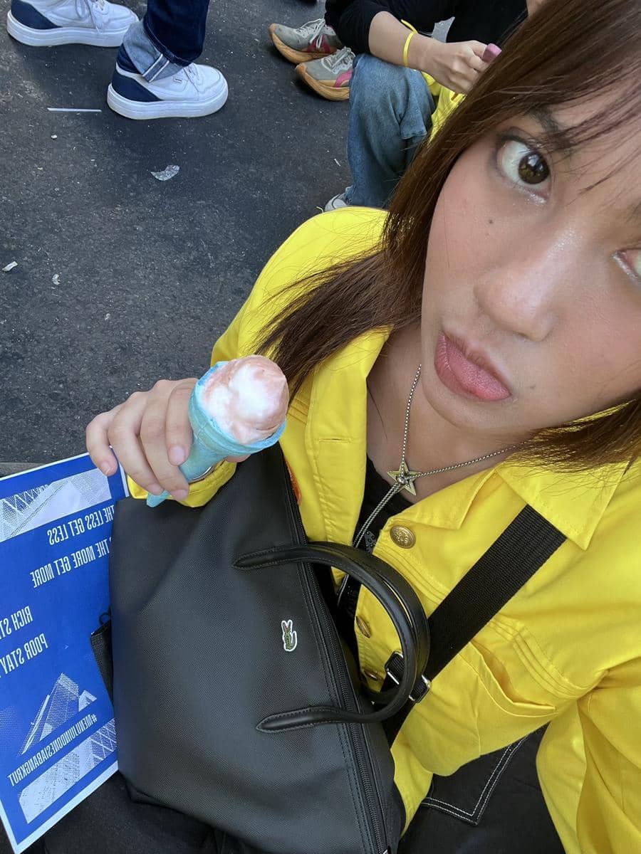 Seorang perempuan mengenakan jaket kuning memegang es krim dan tas hitam saat duduk di jalan bersama beberapa orang lain di sekitarnya.