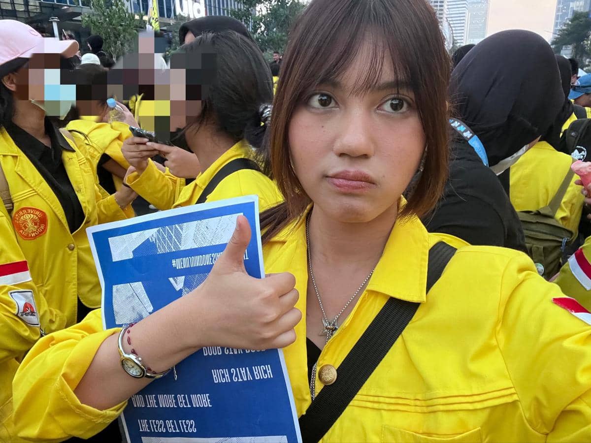 Seorang mahasiswa mengenakan jaket almamater kuning memegang poster dan mengacungkan jempol saat mengikuti aksi demonstrasi bersama rekan-rekannya.