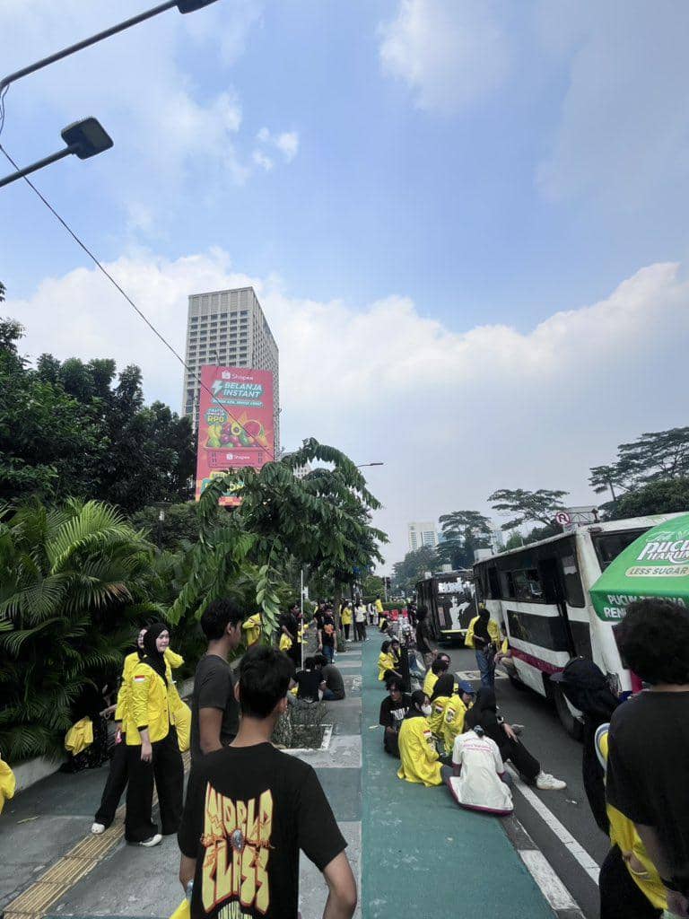 Sejumlah mahasiswa mengenakan jaket kuning berkumpul di trotoar dan jalan kota saat melakukan aksi demonstrasi di bawah langit cerah.