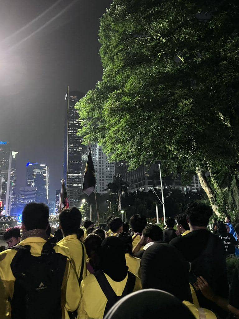 Sekelompok mahasiswa mengenakan jaket kuning berkumpul pada malam hari di area perkotaan dengan gedung tinggi dan pepohonan di sekitarnya.
