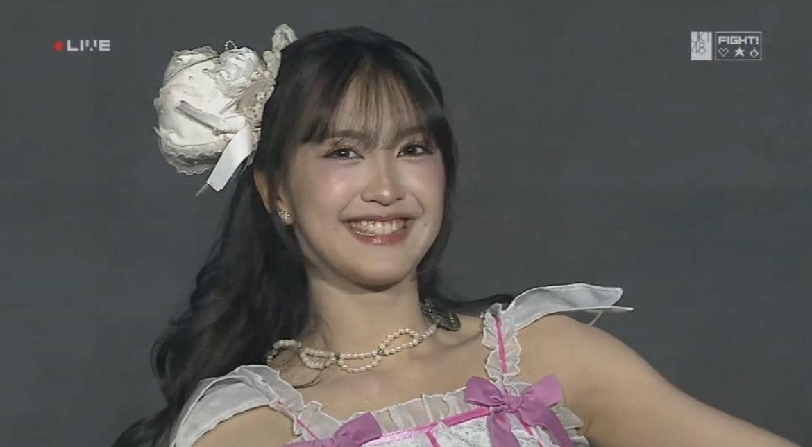 Seorang anggota JKT48 tersenyum di atas panggung saat acara JKT48 Request Hour 2026 dengan busana bernuansa putih dan pita ungu muda.