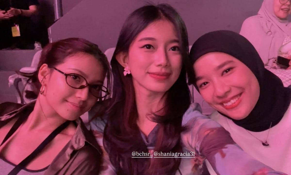 Tiga perempuan berpose bersama sambil tersenyum di sebuah acara dengan pencahayaan ungu lembut dan suasana santai di tribun penonton.