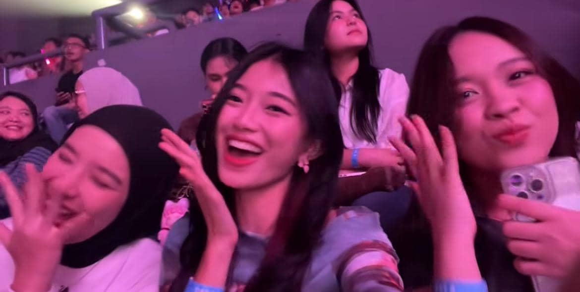Tiga perempuan tersenyum dan berpose ceria di antara penonton konser dengan pencahayaan ungu di area tribun.