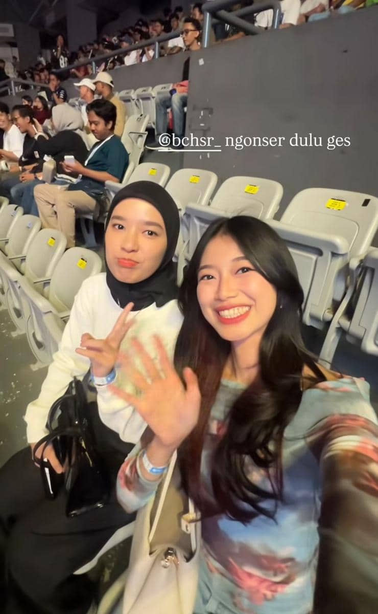 Dua perempuan berpose sambil tersenyum di kursi penonton sebuah acara musik dengan latar penonton lain di tribun.