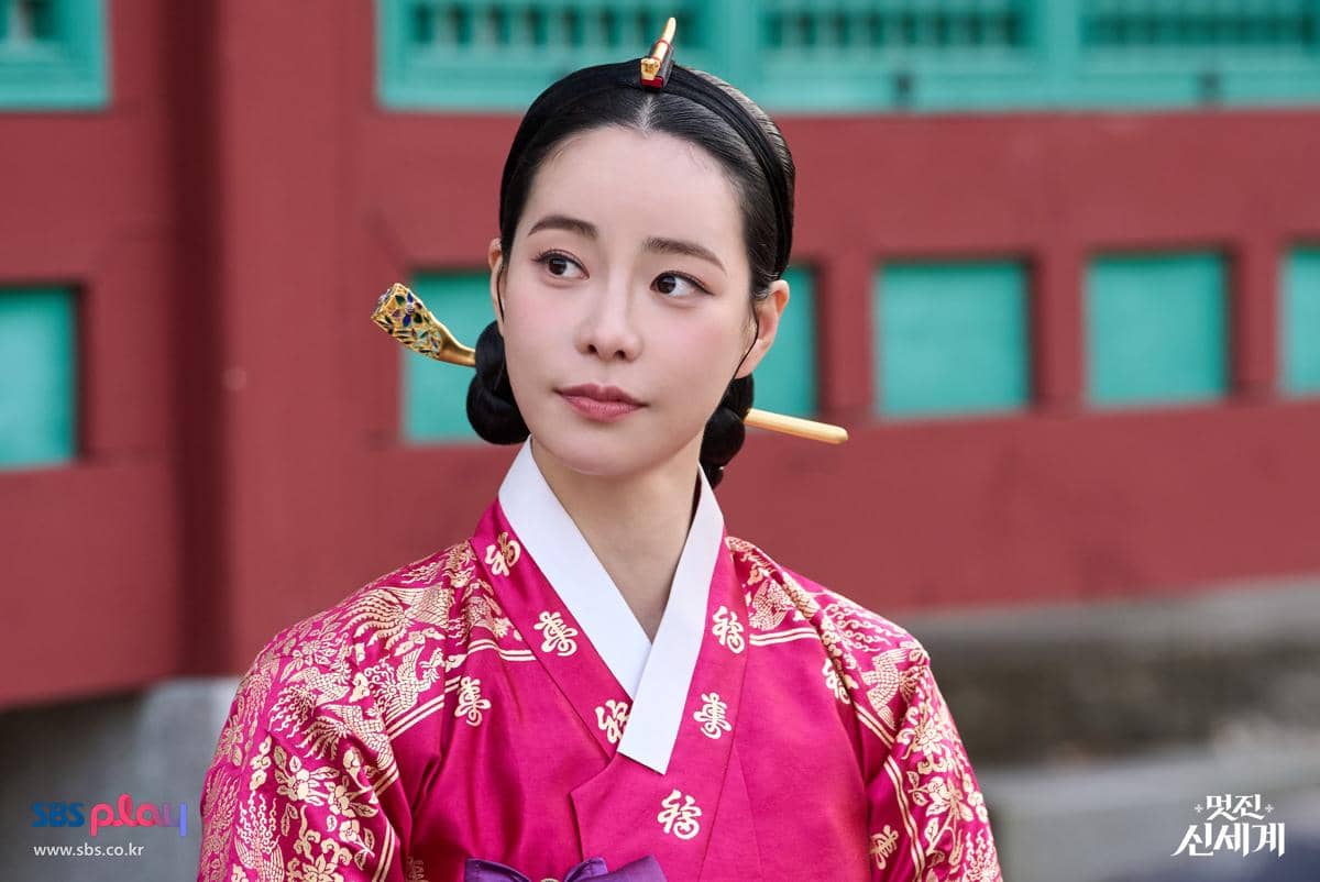 Seorang wanita mengenakan hanbok merah muda berhias motif emas dan aksesori rambut tradisional Korea di latar istana.
