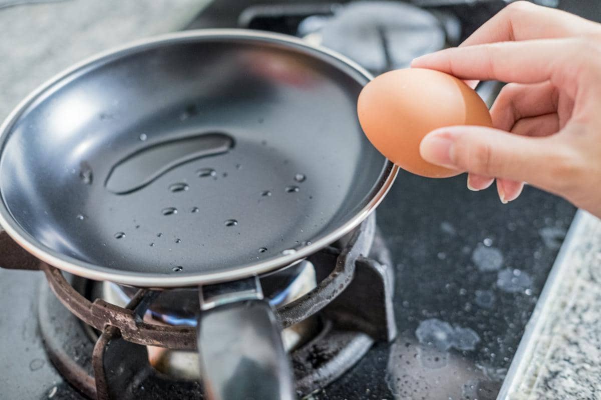 Tangan memegang telur di atas wajan berisi minyak panas di atas kompor gas, persiapan memasak gyeran bap.