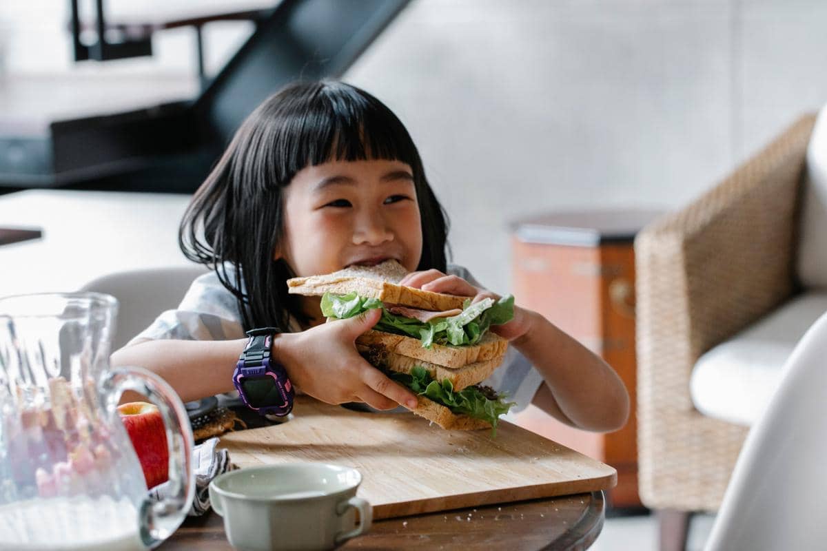 Seorang anak perempuan tersenyum sambil memegang dan menggigit roti lapis besar berisi sayuran di meja makan.