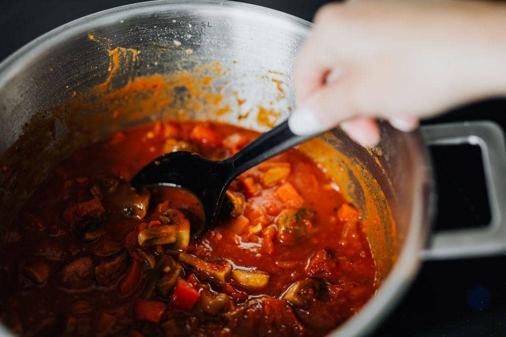 Tangan mengaduk semur berwarna merah dengan sayuran dan potongan daging di dalam panci logam di atas kompor.