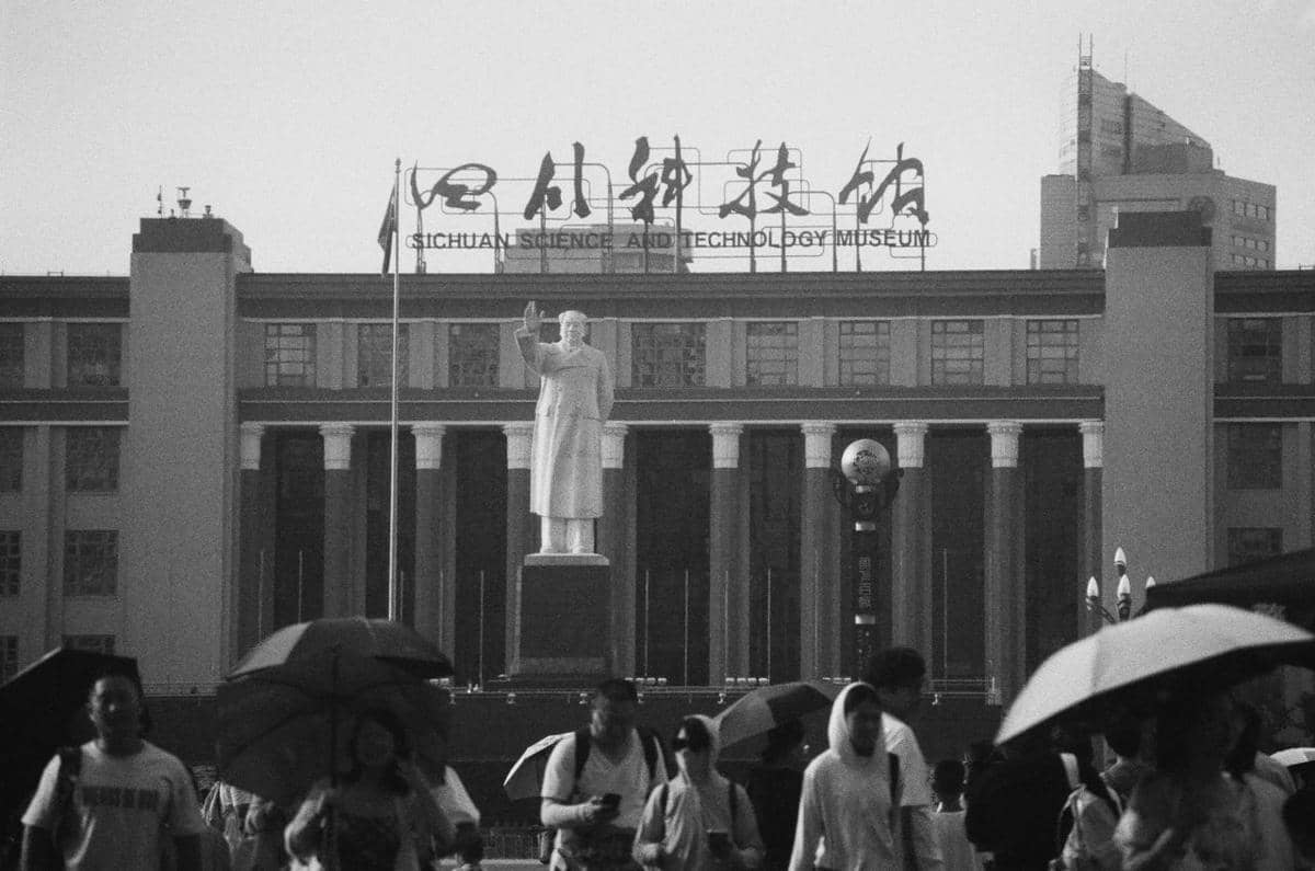 Bangunan Sichuan Science and Technology Museum dengan patung besar di depannya dan orang-orang berjalan sambil membawa payung.