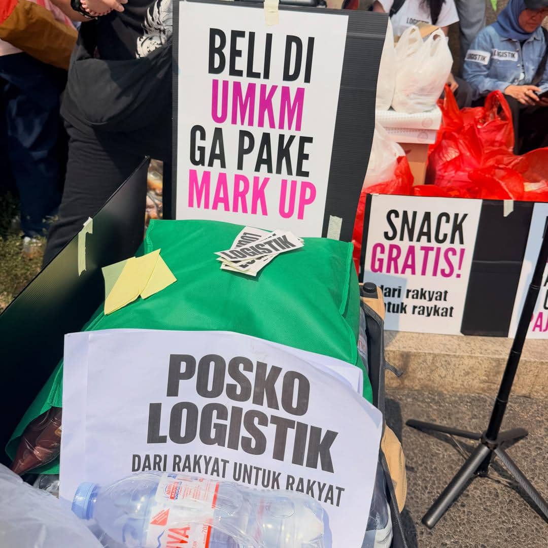Poster bertuliskan pesan dukungan untuk UMKM dan posko logistik dengan tumpukan barang serta botol air di area aksi mahasiswa.