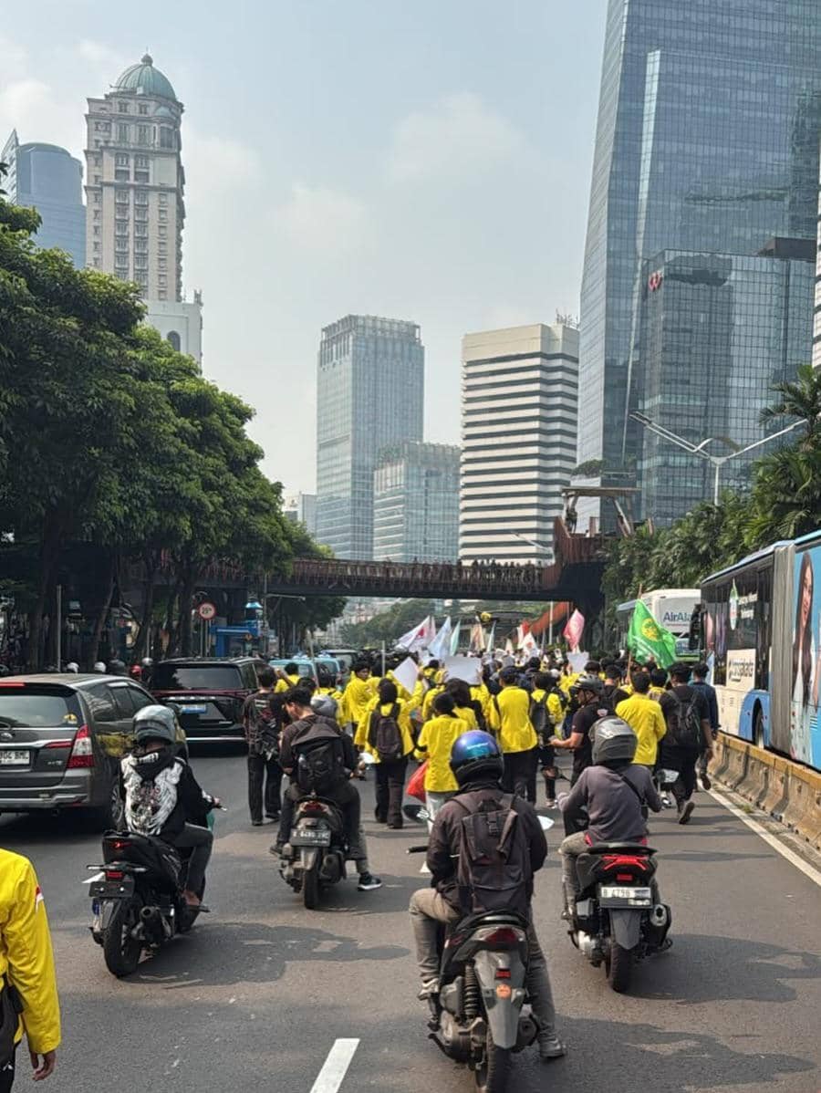 Sejumlah mahasiswa mengenakan jaket kuning berjalan di jalan raya sambil membawa spanduk dan bendera di tengah gedung-gedung tinggi kota.