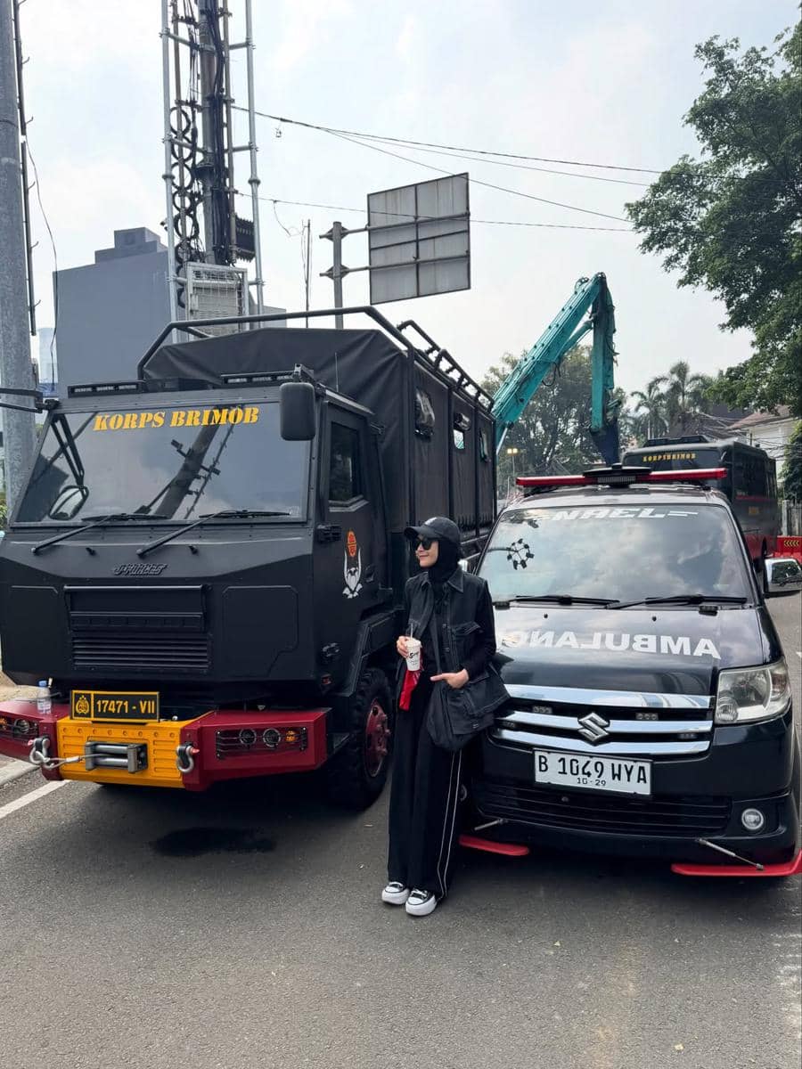 Seorang perempuan berbusana hitam berdiri di antara truk Korps Brimob dan mobil ambulans di tepi jalan kota pada siang hari.