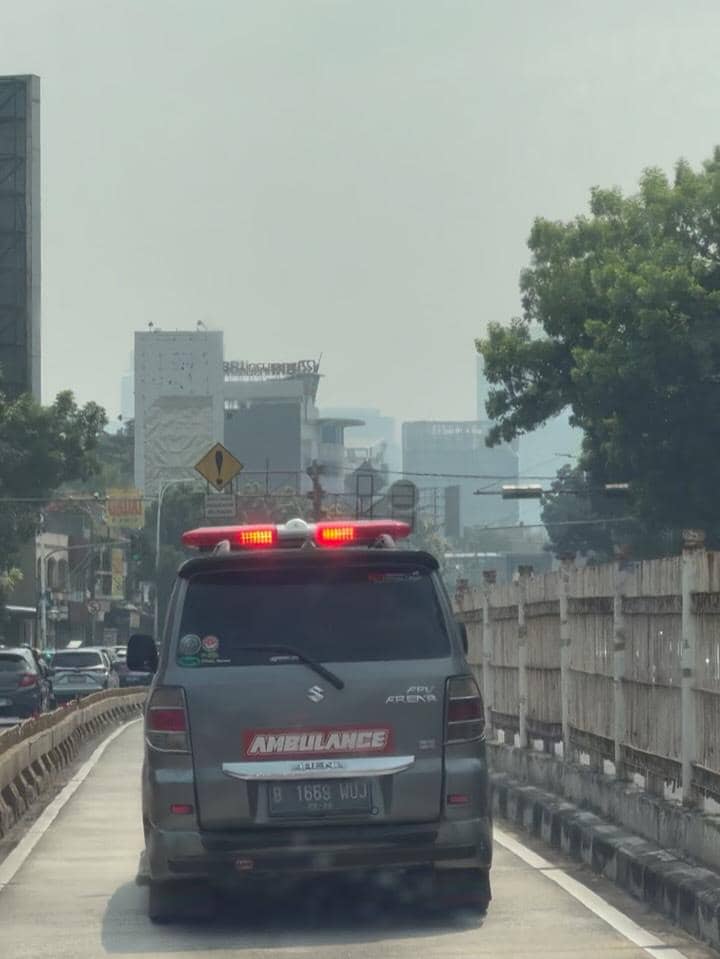 Sebuah mobil ambulans berwarna abu-abu dengan lampu sirene menyala melaju di jalan kota yang ramai di siang hari.