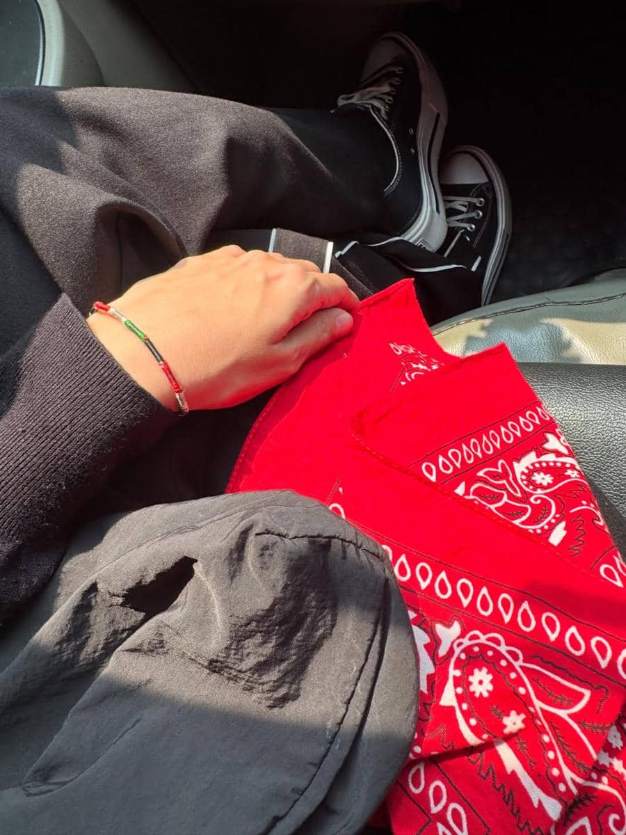 Tangan seseorang mengenakan gelang warna merah, hitam, dan hijau sambil memegang kain bandana merah di atas pangkuan dengan sepatu hitam putih.