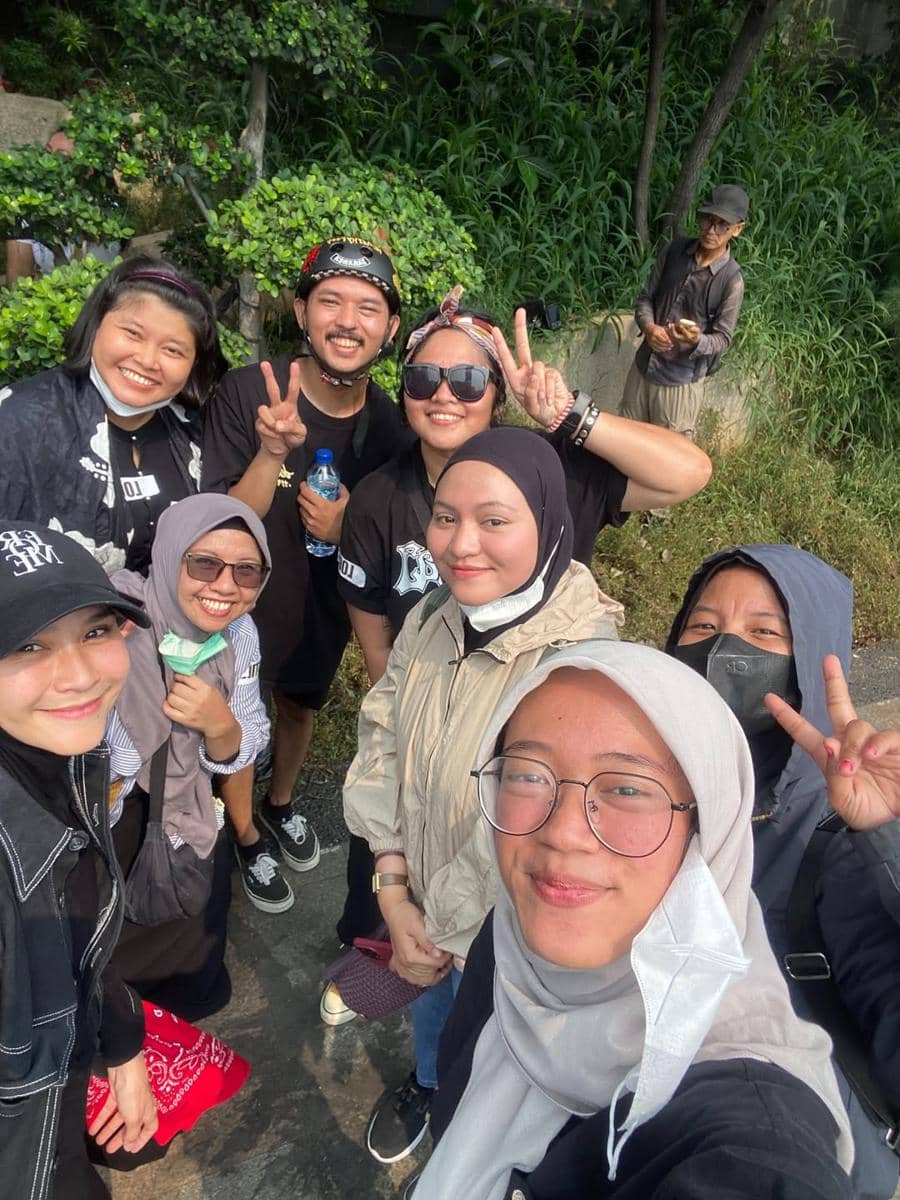 Sekelompok relawan tersenyum dan berpose bersama di luar ruangan dengan latar pepohonan dan semak hijau.