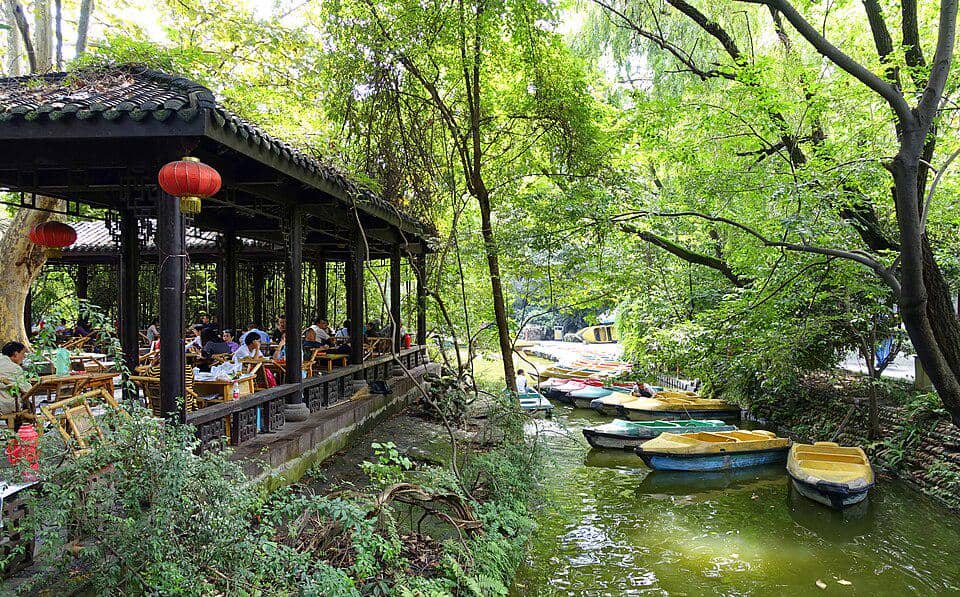 Pemandangan taman dengan paviliun beratap tradisional berisi orang-orang duduk di kursi bambu di tepi sungai kecil dengan perahu warna-warni.
