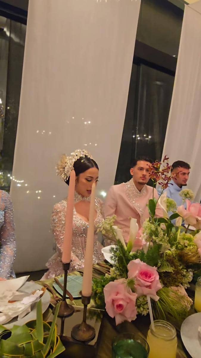 Pasangan pengantin duduk di meja makan malam pernikahan dengan dekorasi bunga mawar merah muda dan lilin berwarna pastel.