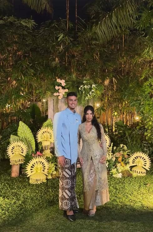 Seorang pria dan wanita berpose bersama di taman malam hari dengan dekorasi bunga dan daun tropis bernuansa tradisional.
