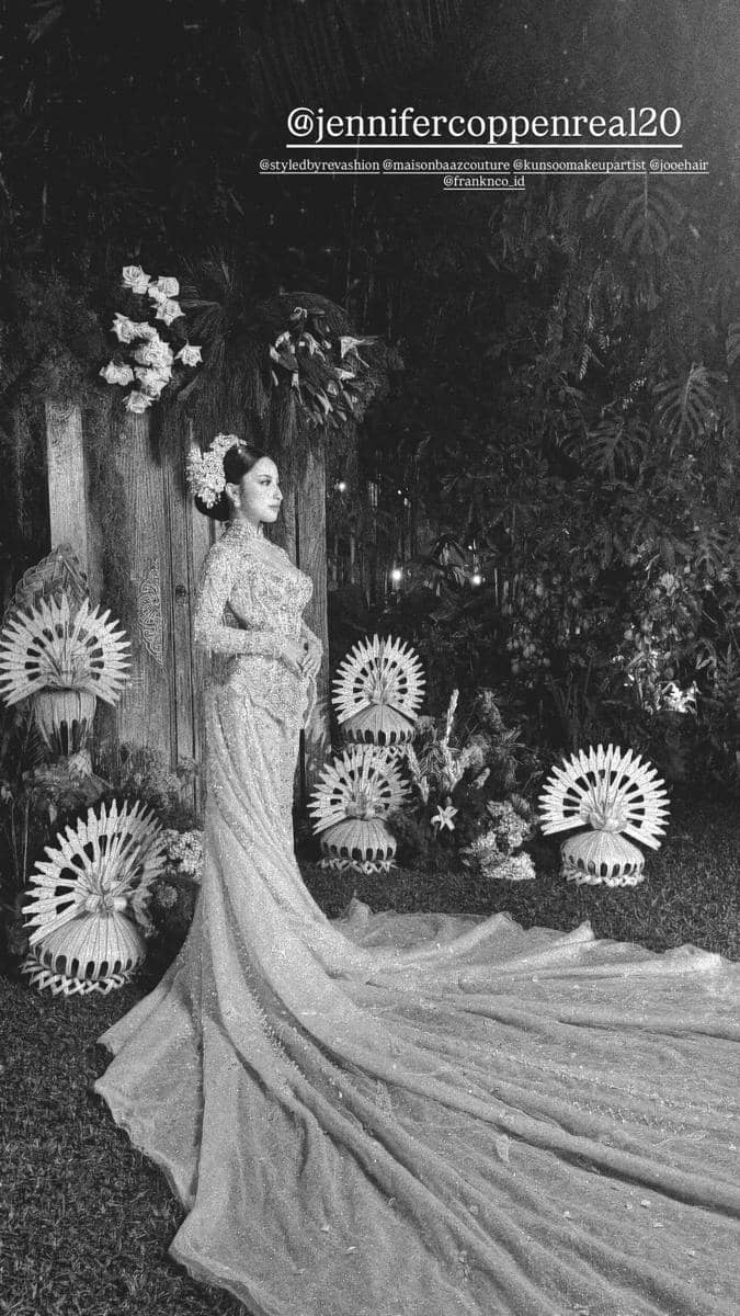 Seorang wanita mengenakan gaun pengantin panjang berhias payet berdiri anggun di taman dengan dekorasi bunga dan ornamen tradisional.