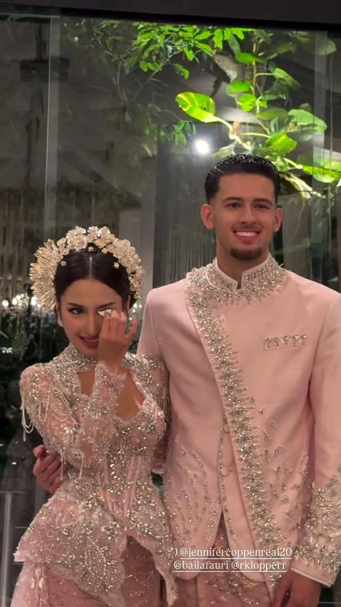 Sepasang pengantin mengenakan busana adat modern berwarna pink berhias payet dan ornamen perak saat acara resepsi pernikahan.