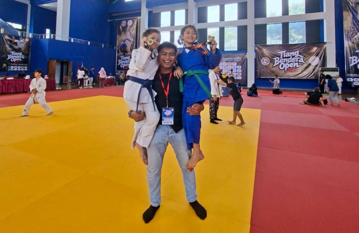 Dua atlet muda judo mengenakan gi putih dan biru berpose gembira bersama pelatih di atas matras usai meraih medali emas di Tiang Bendera Open 2026.