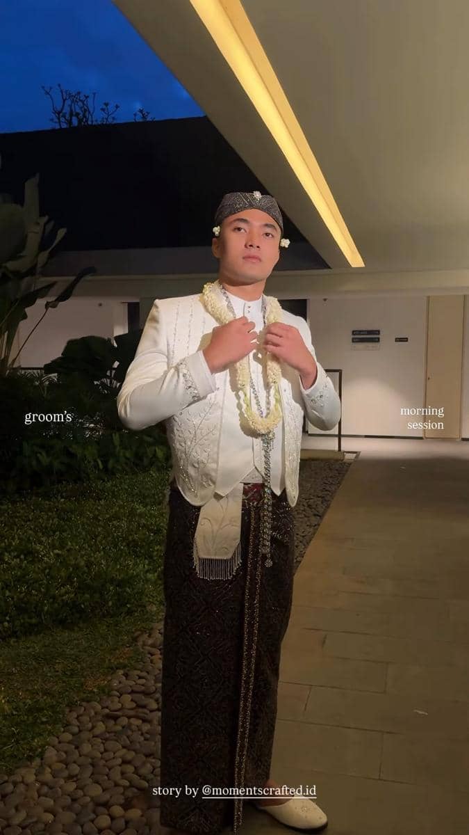 Seorang pria mengenakan busana pengantin adat Sunda berwarna putih dengan blangkon dan kalung melati di area luar ruangan saat sesi pagi.