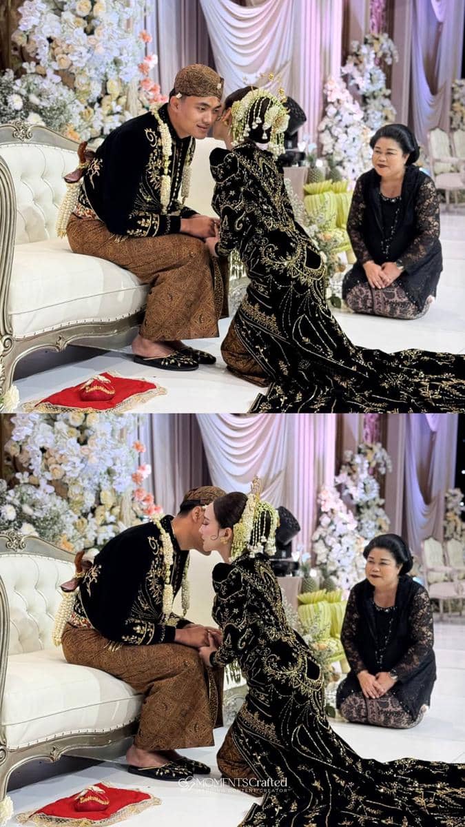 Pasangan pengantin mengenakan busana adat Jawa berwarna hitam dan emas dalam prosesi pernikahan di pelaminan berhias bunga putih.