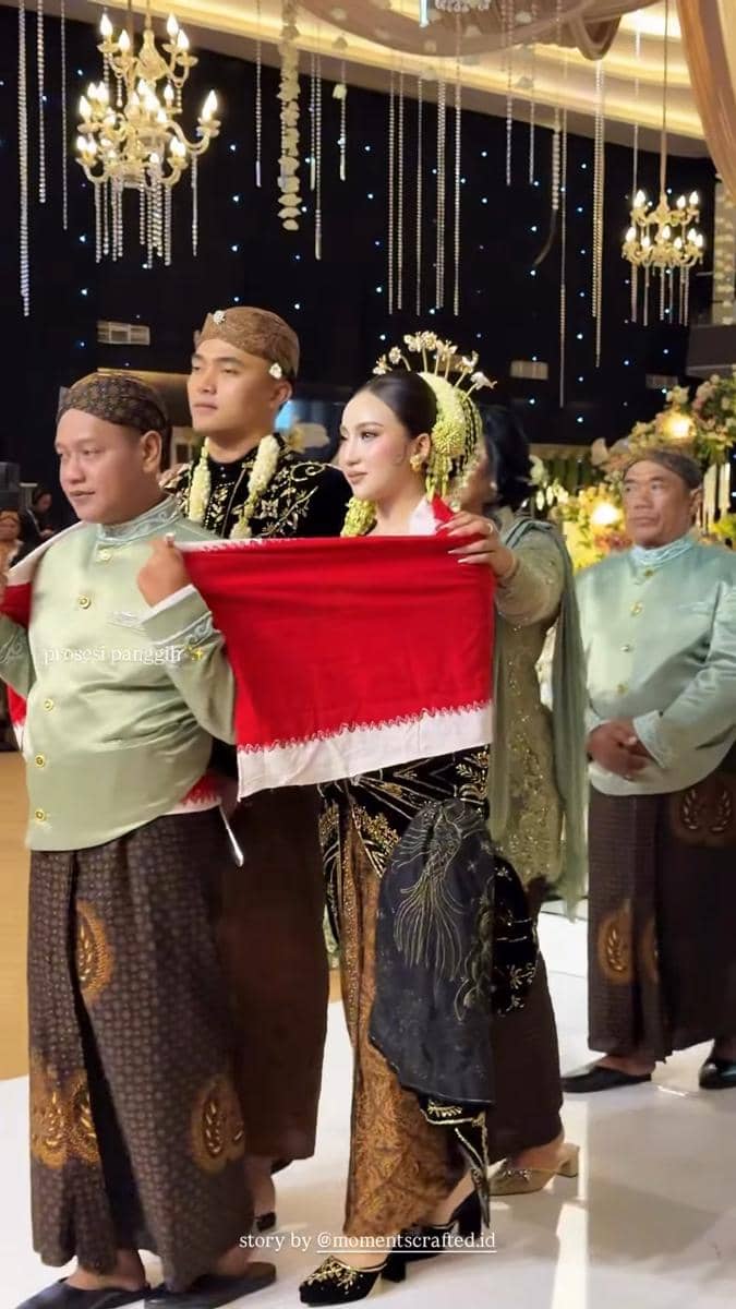 Pasangan pengantin mengenakan busana adat Jawa berwarna hitam dan emas saat prosesi pernikahan, memegang kain merah putih di dalam gedung berhias lampu gantung.