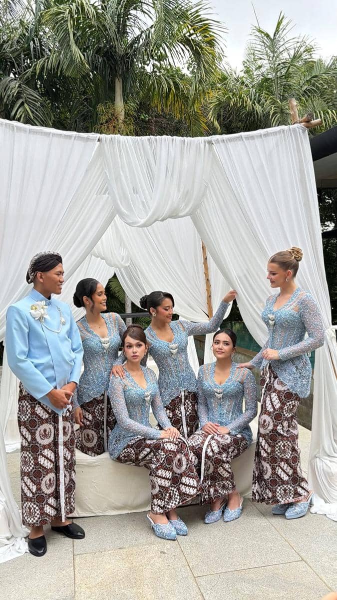 Lima perempuan dan satu pria mengenakan busana tradisional kebaya biru dan kain batik berpose di bawah tirai putih di acara akad.