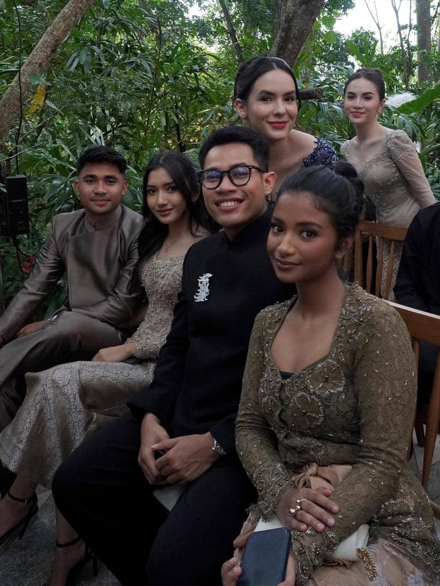 Sekelompok tamu berpakaian formal duduk bersama di acara akad pernikahan dengan latar pepohonan hijau di luar ruangan.