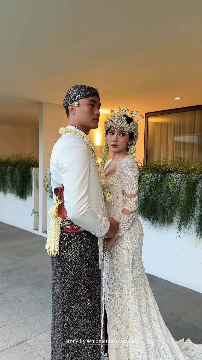 Pasangan pengantin mengenakan busana adat Jawa, mempelai pria memakai beskap putih dan blangkon, sedangkan mempelai wanita mengenakan kebaya putih berhias siger dan ronce melati.