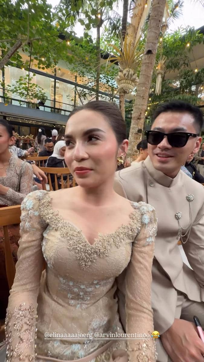 Seorang wanita mengenakan kebaya modern berwarna krem duduk di samping pria berkacamata hitam di acara akad pernikahan outdoor.