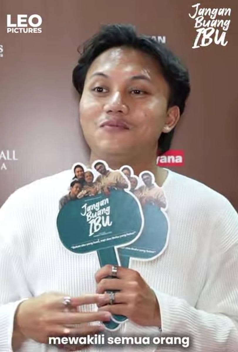 Seorang pria mengenakan sweater putih memegang kipas promosi bertuliskan 'Jangan Buang Ibu' di acara peluncuran film.