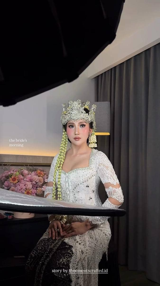Seorang pengantin wanita mengenakan kebaya putih berhias mahkota dan ronce melati sedang duduk di ruang rias dengan latar bunga dan tirai abu-abu.