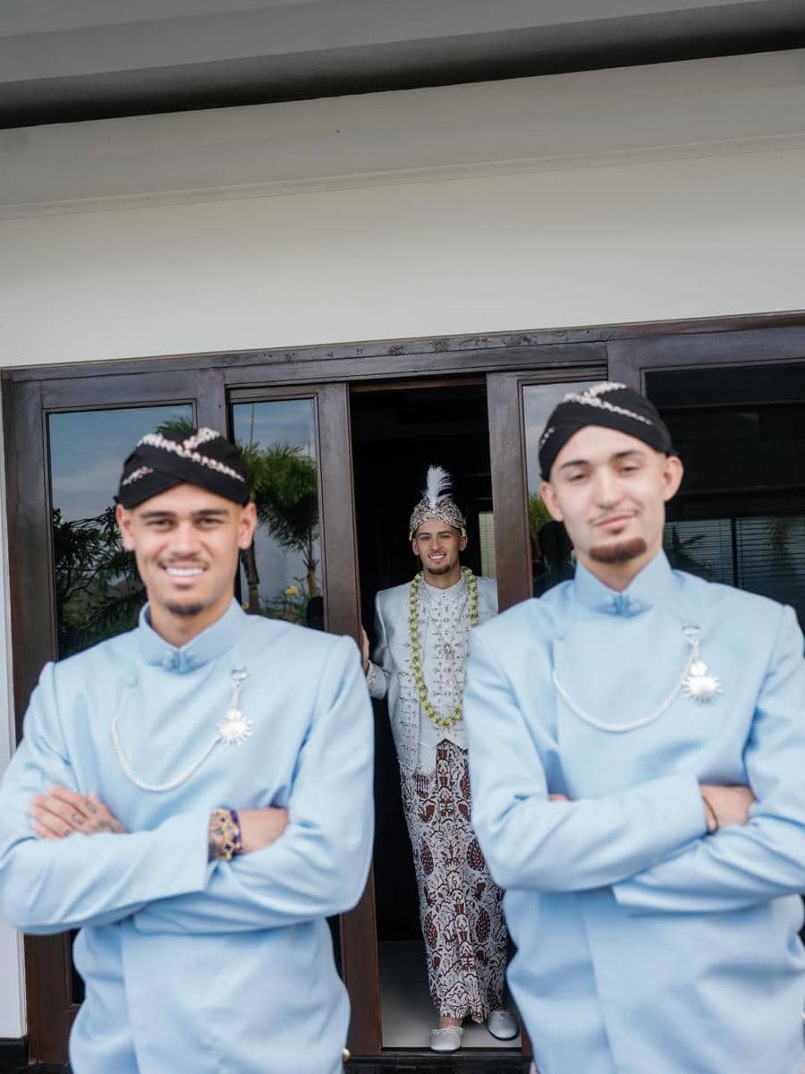 Dua pria mengenakan beskap biru muda berdiri di depan pintu, sementara seorang pria berpakaian adat pengantin Jawa berdiri di belakang mereka.