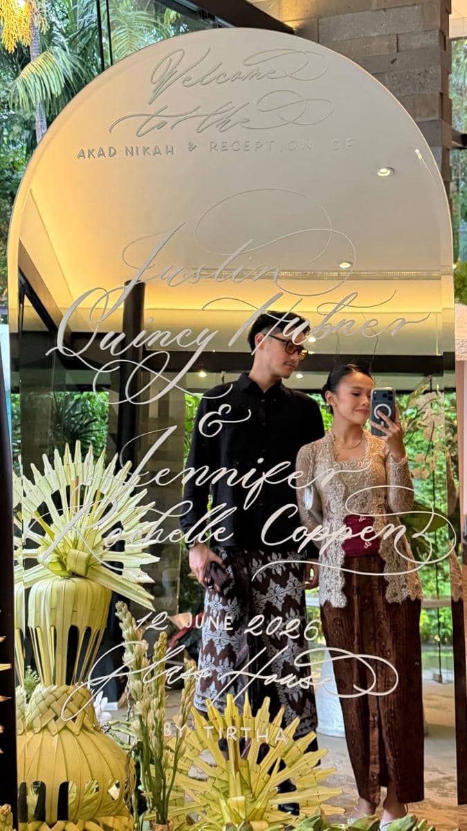Sepasang tamu berpakaian tradisional menghadiri akad nikah Jennifer Coppen dan Justin Hubner, berpose di depan cermin dekoratif bertulisan nama pengantin.