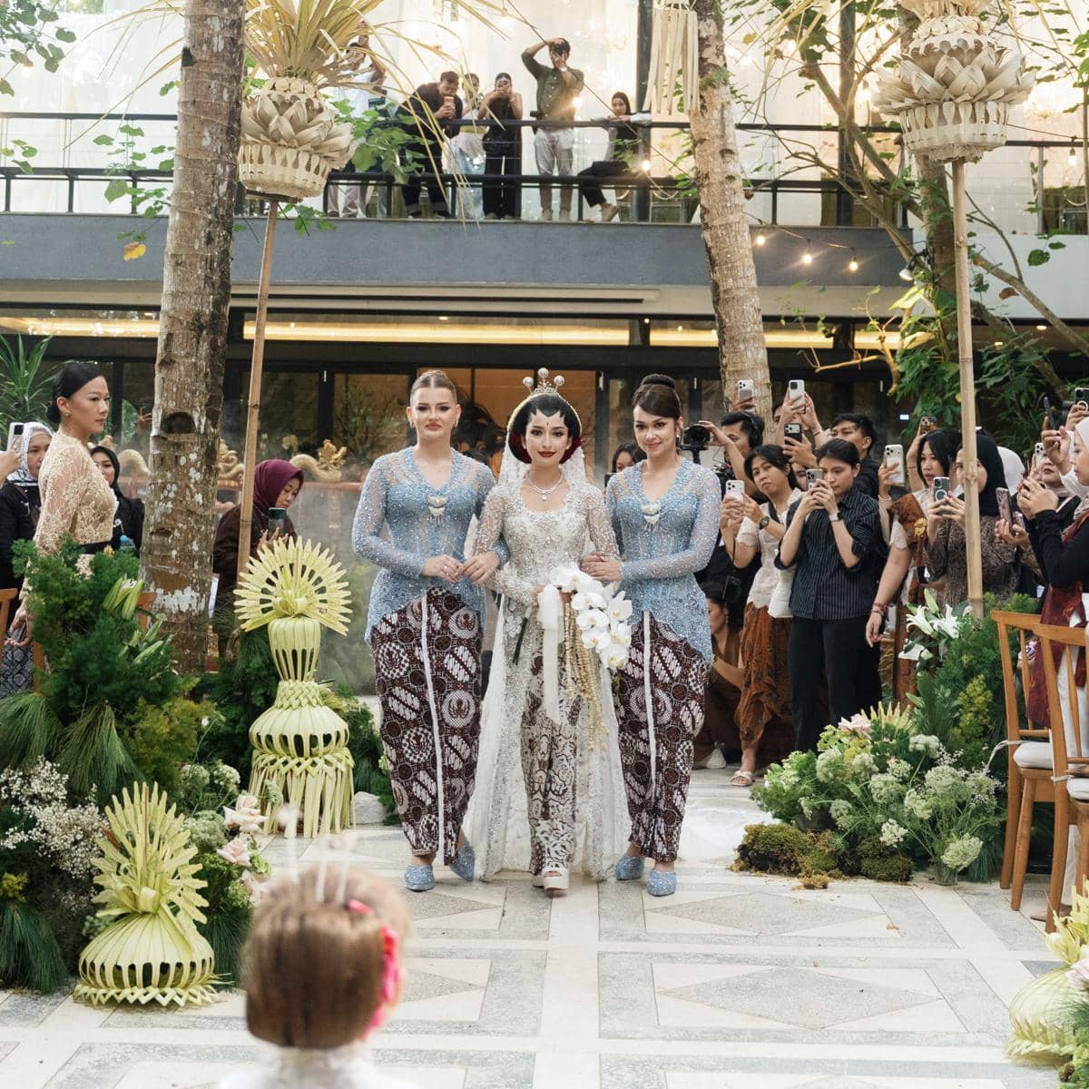 Pengantin wanita berjalan diapit dua bridesmaid mengenakan kebaya biru dan kain batik dalam prosesi akad dengan dekorasi adat Jawa.