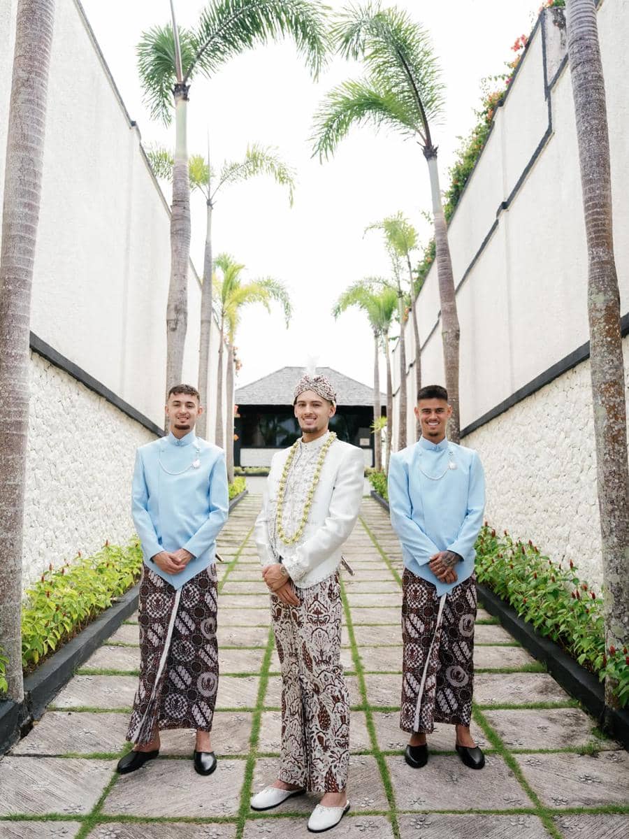 Tiga pria mengenakan busana adat Jawa dengan beskap dan kain batik berdiri di jalan berpohon palem di area luar ruangan.