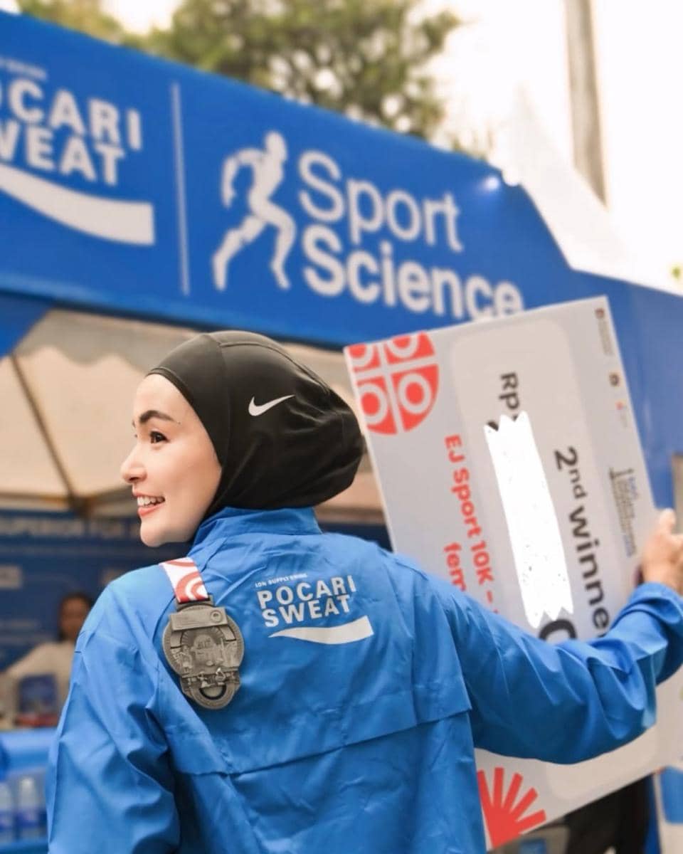 Seorang peserta wanita berhijab mengenakan jaket biru Pocari Sweat tersenyum sambil memegang papan hadiah di ajang lari Jakarta International Marathon 2026.