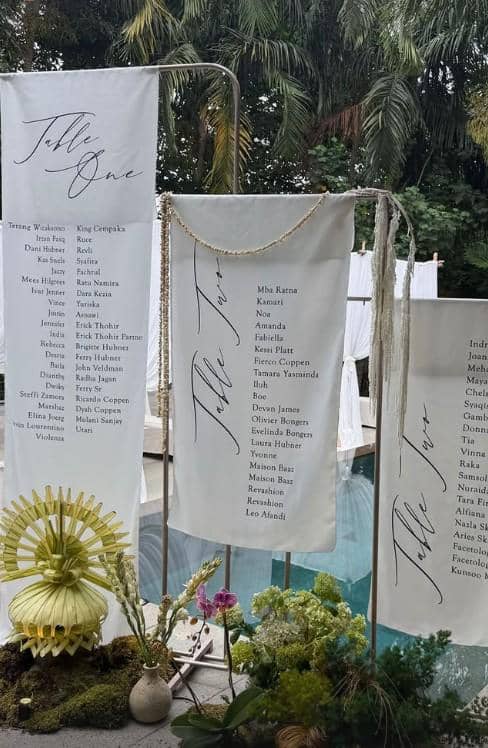 Dekorasi daftar meja tamu pernikahan bergaya elegan dengan kain putih bertuliskan nama-nama tamu di tepi kolam renang dan hiasan bunga tropis.