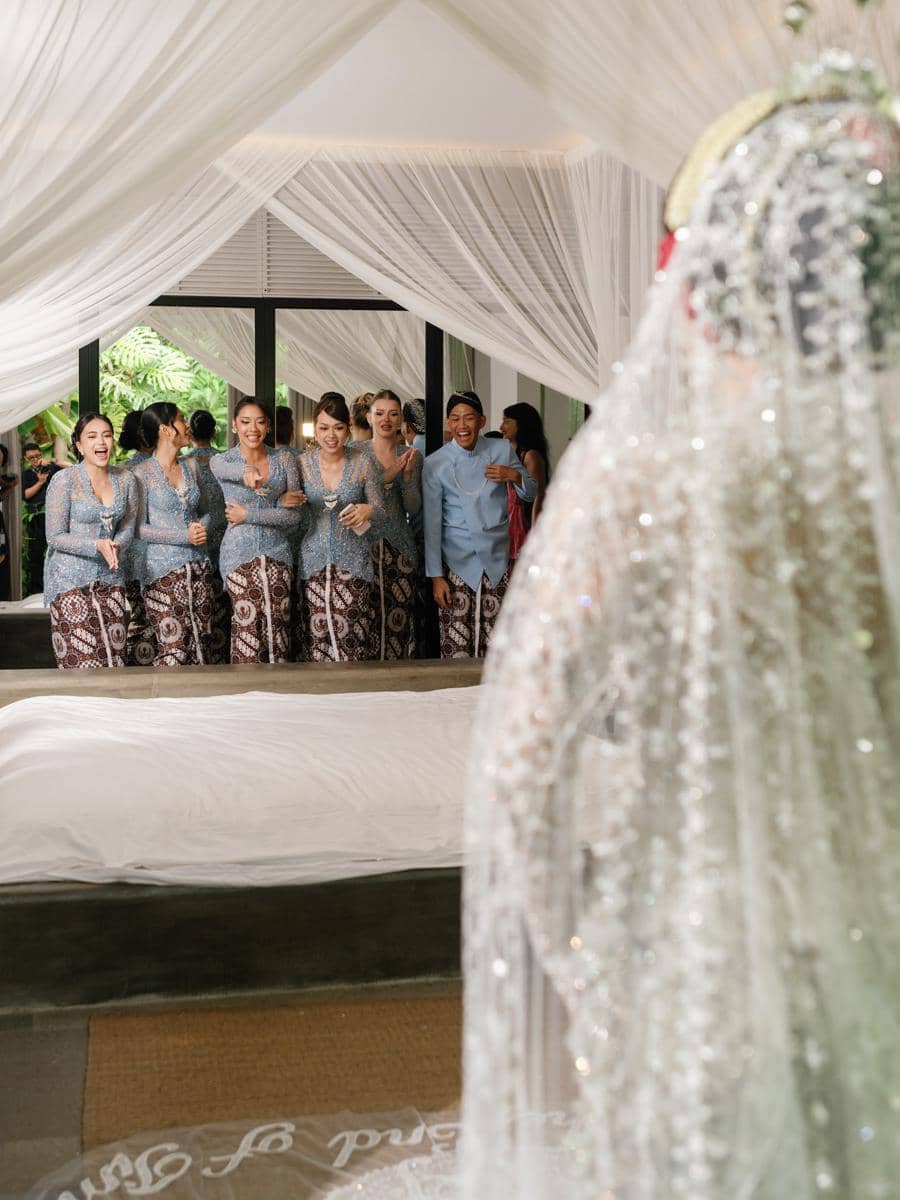 Para bridesmaid mengenakan kebaya biru dan kain batik tersenyum gembira melihat pengantin wanita bersiap untuk akad nikah.