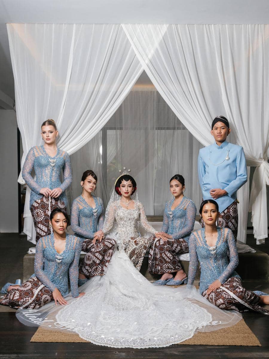 Pengantin wanita duduk di tengah mengenakan kebaya putih dengan enam bridesmaid berkebaya biru dan kain batik di ruangan berhias tirai putih.