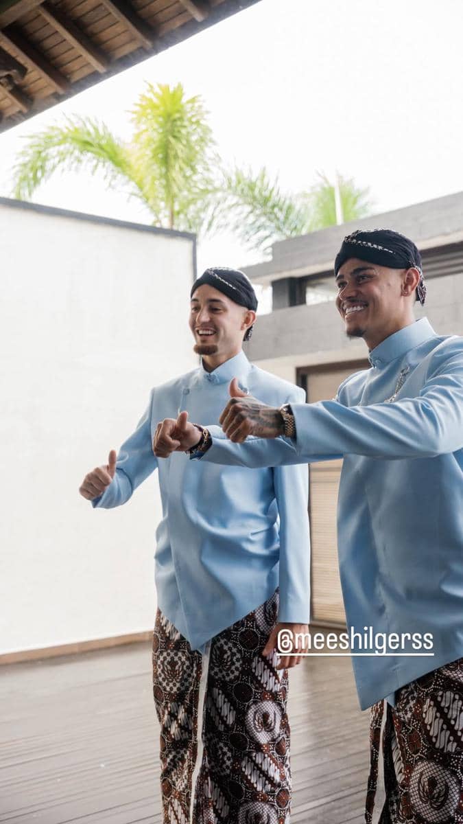 Dua pria mengenakan beskap biru muda dan kain batik lengkap dengan blangkon, tersenyum sambil berpose di area terbuka.
