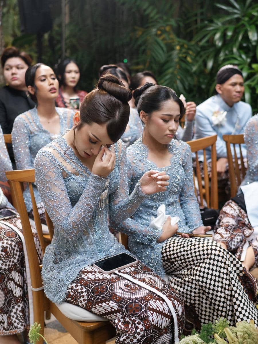 Para bridesmaid mengenakan kebaya biru dan kain batik duduk di acara akad, dengan salah satu perempuan menyeka air mata haru.